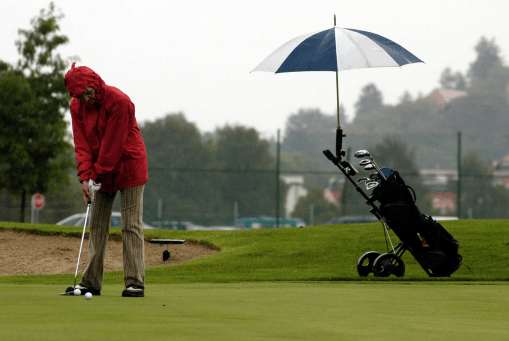 golfer playing in the rain wearing a raincoat with an umbrella protecting his clubs